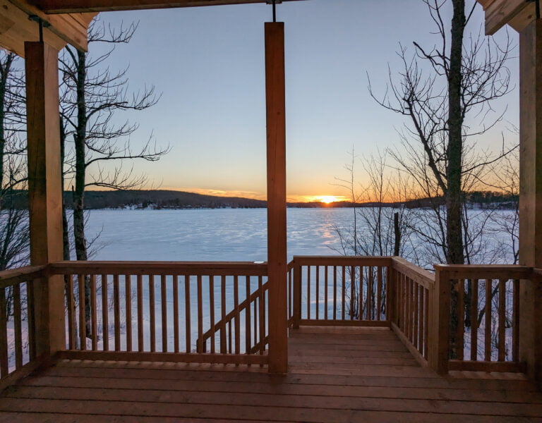 A stunning view of the frozen lake and the setting sun from the porch of a cabin.