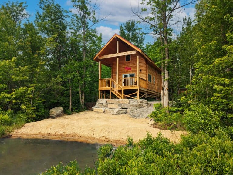 A quaint wooden cabin sits above a small, sandy beach that gently slopes into a calm pond. Surrounded by lush greenery, the beach offers a perfect spot for wading, relaxing, or enjoying nature's peace.