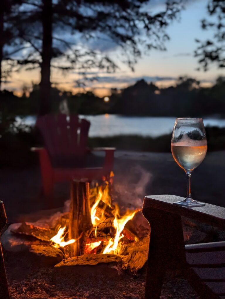 A glowing campfire burns brightly in a stone fire pit, surrounded by red Adirondack chairs. The scene is set at dusk with silhouettes of trees and a calm lake in the background, under a darkening sky.