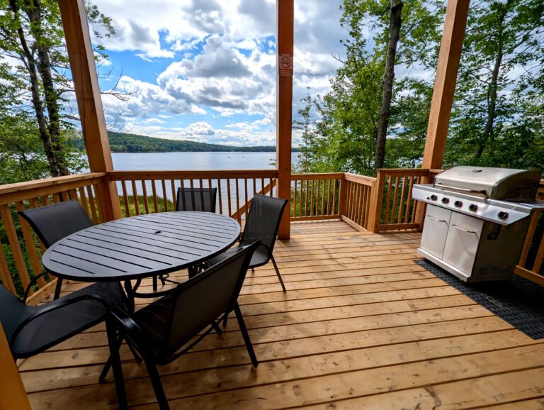 A wooden deck overlooking a calm lake, featuring a round black table with mesh chairs. A gas grill sits nearby framed in sunlight. The deck is framed by tall trees, offering a peaceful spot with a picturesque view.