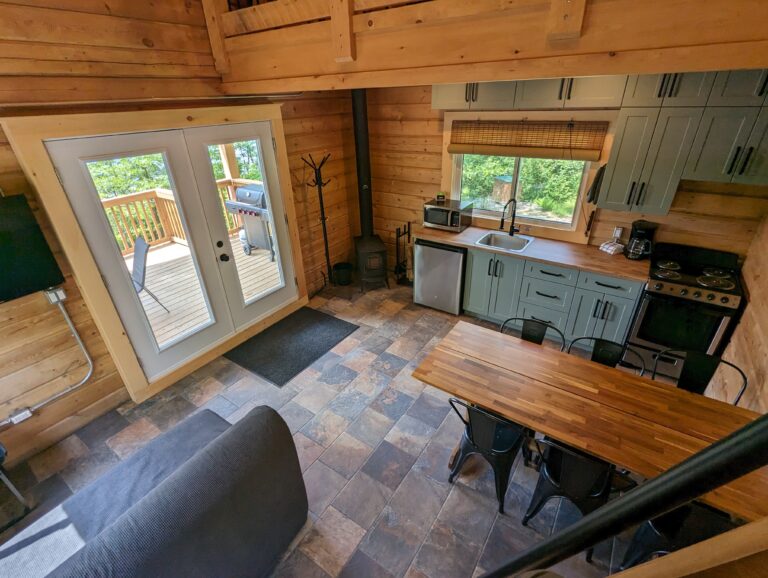 Open living space with a view of the kitchen, dining area, and glass doors leading to a deck with a grill. Modern kitchen with teal cabinets and wood countertops. Stone tile flooring throughout, wood stove in the corner.