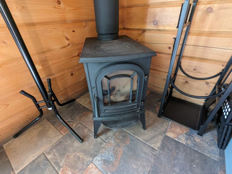 A small corner with a black cast-iron wood stove on stone tile flooring, bordered by wood-paneled walls. Fire tools and a wood holder are neatly arranged beside the stove.