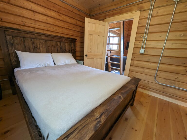 Cozy bedroom with a rustic wooden bed frame and a comfortable queen-sized bed. Wood-paneled walls and natural wood flooring enhance the cabin feel. Doorway opens to the main living area with a visible space-saving staircase.