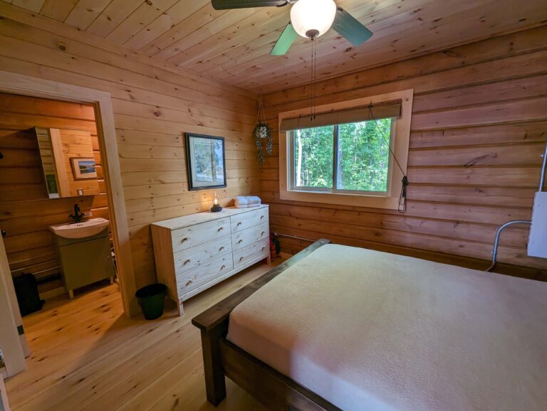 Bright bedroom with wood-paneled walls and ceiling, featuring a large window, ceiling fan, wooden dresser, and attached bathroom. Natural light enhances the cozy, rustic atmosphere. Photo from the perspective of entering the room.