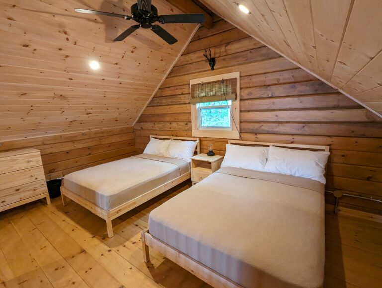 Loft bedroom with two double-sized beds, wood-paneled walls and ceiling, ceiling fan, and a small window. A wooden dresser and nightstand add storage. Soft lighting inset creates a cozy, rustic atmosphere.