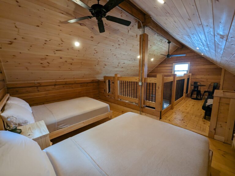 Spacious loft with two double beds, wood-paneled walls and ceiling, and ceiling fans. Open railing overlooks the lower level, with a cozy seating area and a window providing natural light.