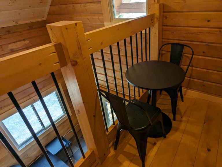 Small loft seating area with a black round table and two matching chairs. Wood-paneled walls and railing overlook the lower level. A nearby window brings in natural light, creating a cozy nook.