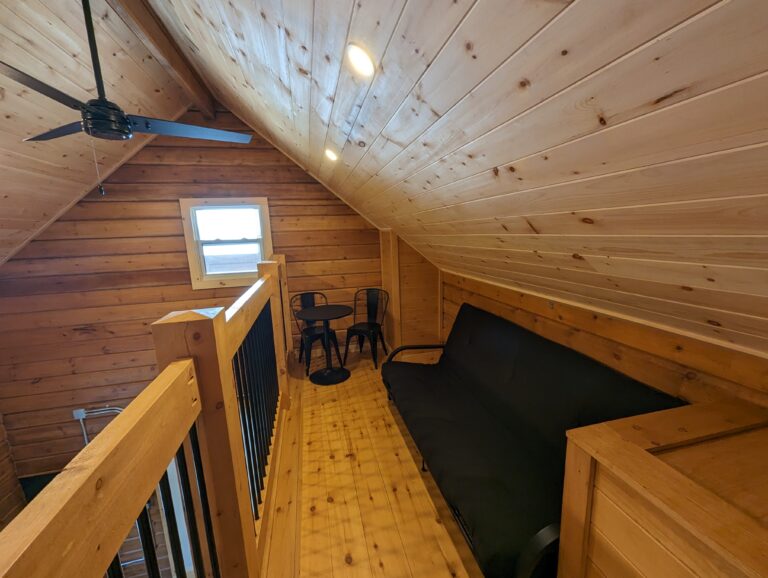 Cozy loft seating area with wood-paneled walls and ceiling, featuring a black futon, small table with two chairs, ceiling fan, and a window. Overlooks the lower level.