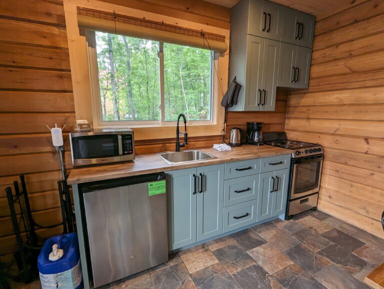 Kitchen with modern appliances, including a stove, microwave, and half-fridge (5.6 cubic ft). Adjacent wooden dining table with seating for six. Large window provides natural light and views of surrounding trees.