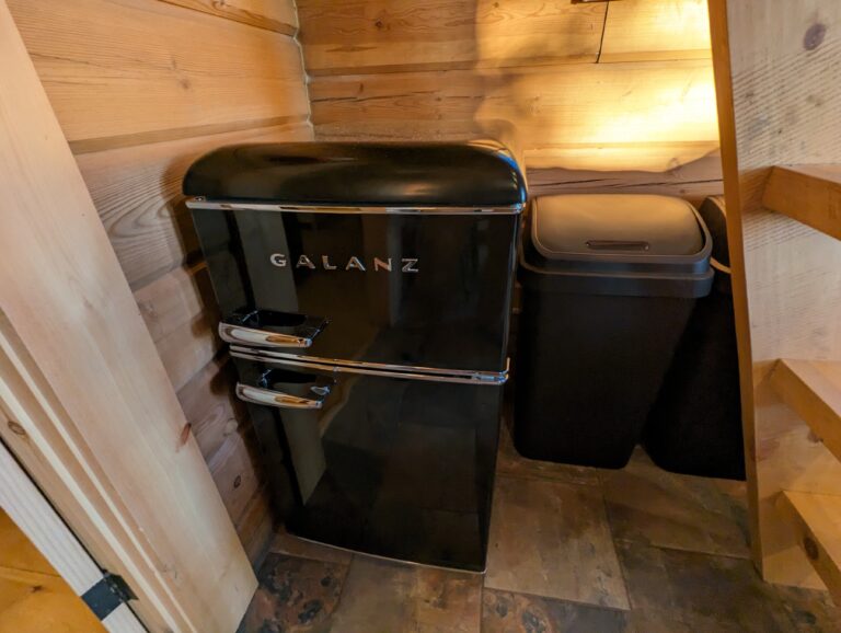 Compact area with a retro-style black Galanz mini-fridge and matching trash bin, tucked in a corner against wood-paneled walls. Located near the stairs
