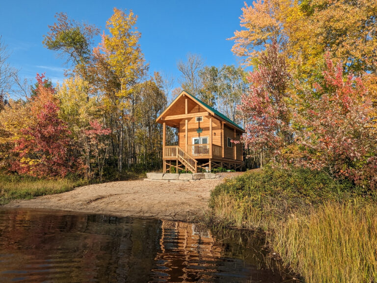 A charming wooden cabin with a covered porch sits directly on a sandy beachfront, surrounded by lush trees. The calm water of the lake laps gently at the shore, creating a peaceful, inviting scene.