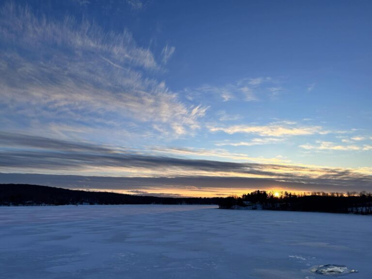 A serene winter scene of a frozen lake under a vast sky. The sunset casts a soft golden glow on the horizon, with delicate clouds stretching across the sky.