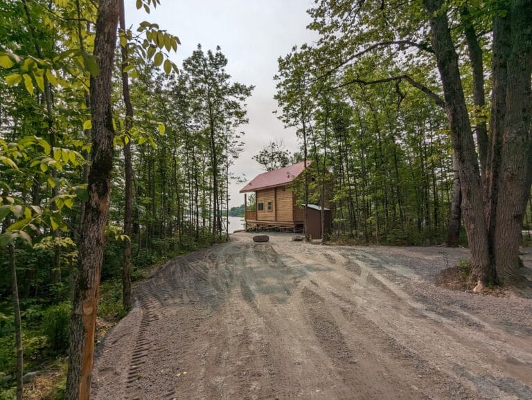 A spacious gravel parking area surrounded by tall trees leads to a wooden cabin with a roof, perched near the lake. Ample room for multiple vehicles, ensuring easy access to this secluded retreat.
