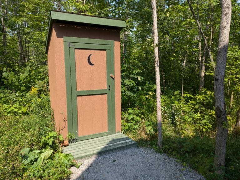 A quaint wooden outhouse stands on a gravel path amidst tall trees. The door features a classic crescent moon cutout, adding rustic charm to this practical amenity nestled in the peaceful woodland setting.