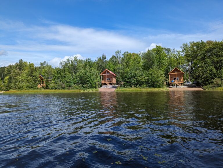 Three cozy wooden cabins stand along a lush, green shoreline, each with its own deck facing a calm lake. The clear blue sky and gentle water create a tranquil setting, perfect for a peaceful retreat in nature. Caption includes further context.